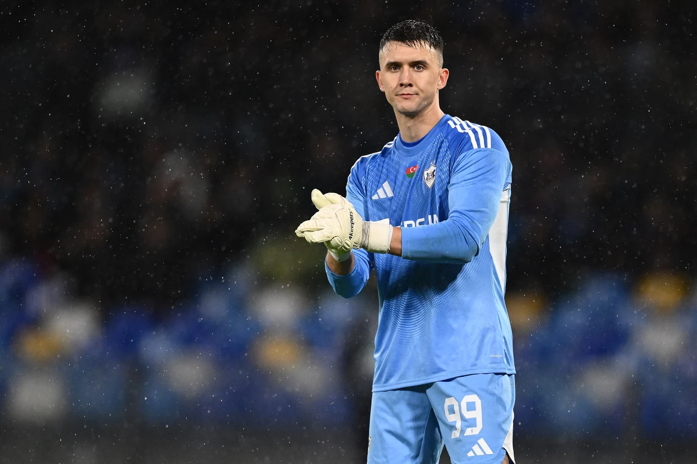 Mateusz Kochalski Bramkarz w niebieskim stroju sportowym stoi na boisku z rękawicami na dłoniach, lekko uśmiechnięty, w tle rozmyte trybuny stadionu.