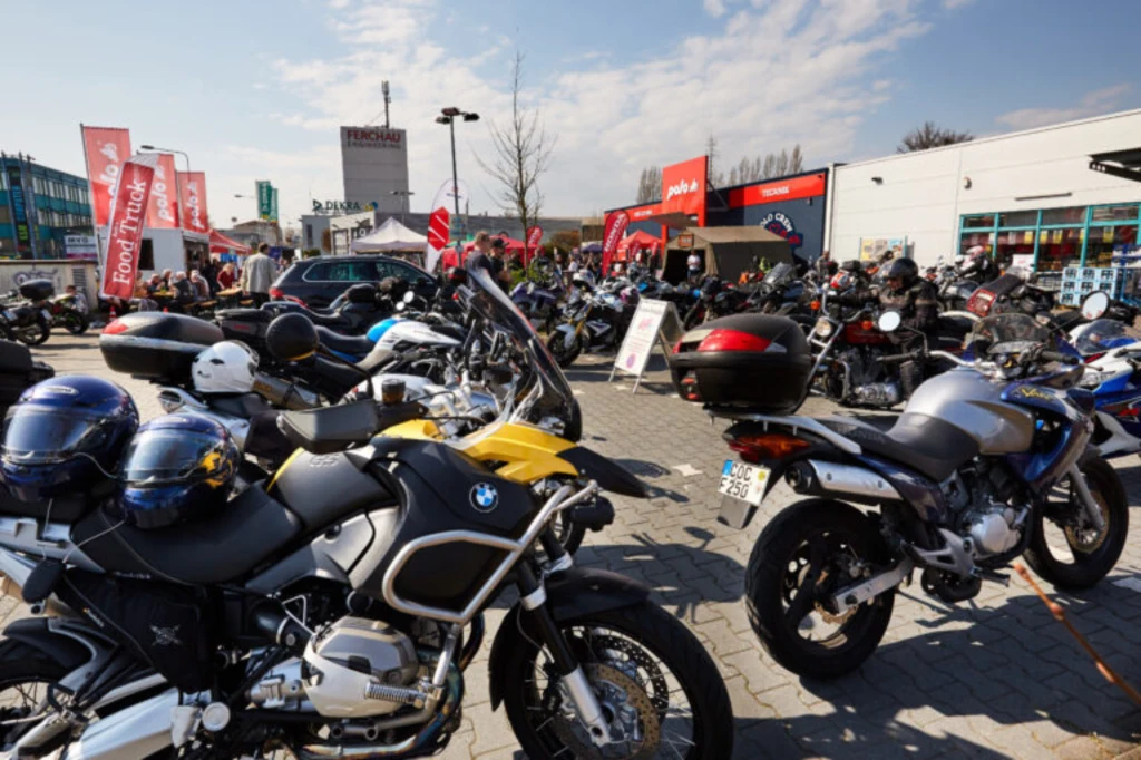 Kilka motocykli zaparkowanych na placu przed sklepami i punktami usługowymi, tłum ludzi w tle, flagi i banery reklamowe sugerujące imprezę motocyklową.