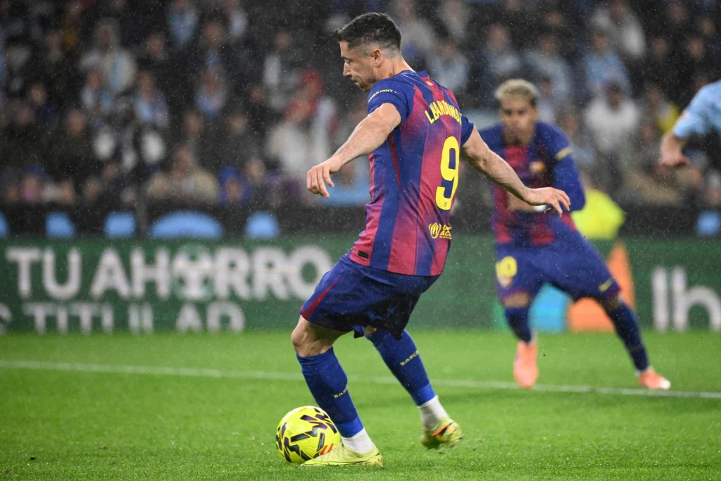 Robert Lewandowski w barwach FC Barcelona w sezonie 2025/2026 Piłkarz w koszulce Barcelony z numerem 9 kopie piłkę podczas meczu, w tle widoczny inny zawodnik oraz rozmyte trybuny.