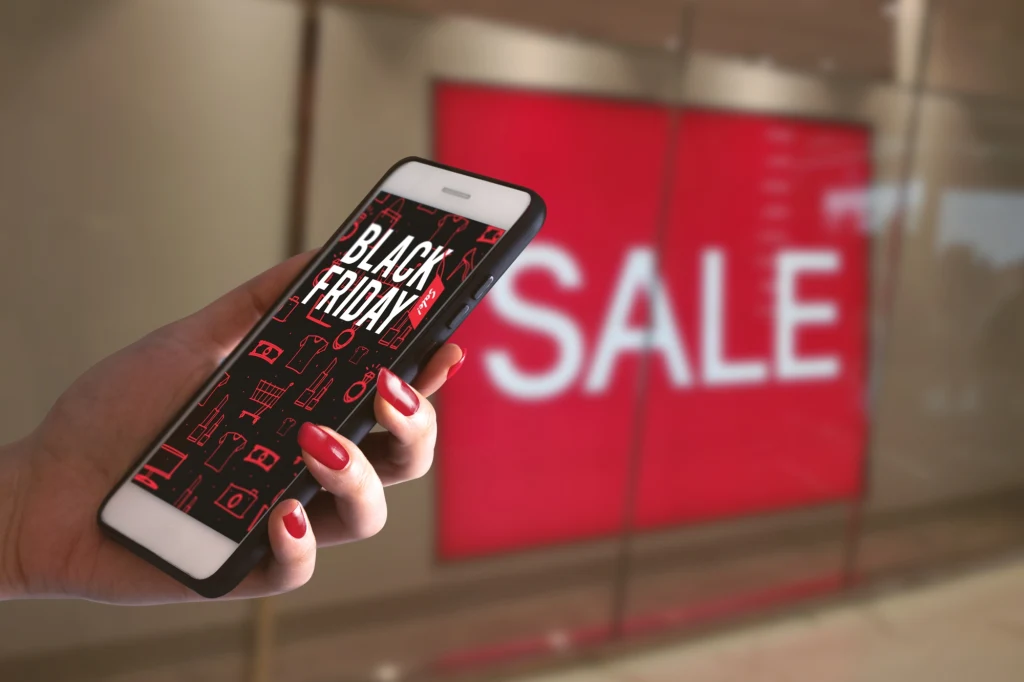 Czy Black Friday to dobra okazja do zakupu nowego smartfona? Ręka trzymająca smartfon z wyświetlonym napisem Black Friday, w tle duży czerwony baner z napisem Sale w witrynie sklepowej.