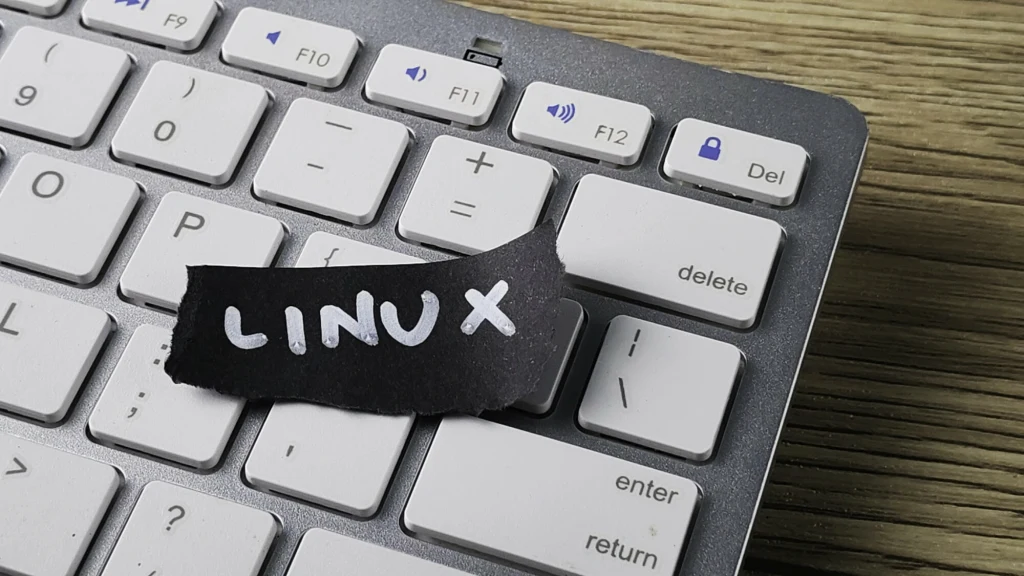 Gaming na Linuxie 2025: Przegląd zmian w Proton 10 Fragment klawiatury komputerowej z naklejonym kawałkiem czarnego papieru z napisem LINUX wykonanym białym markerem, podświetlenie klawiszy oraz drewniane tło.