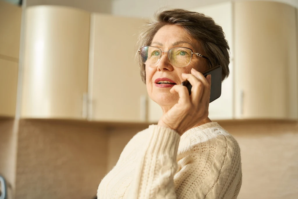 Starsza kobieta w okularach, ubrana w jasny sweter, rozmawia przez telefon komórkowy w kuchni. Tło stanowią jasne szafki kuchenne.