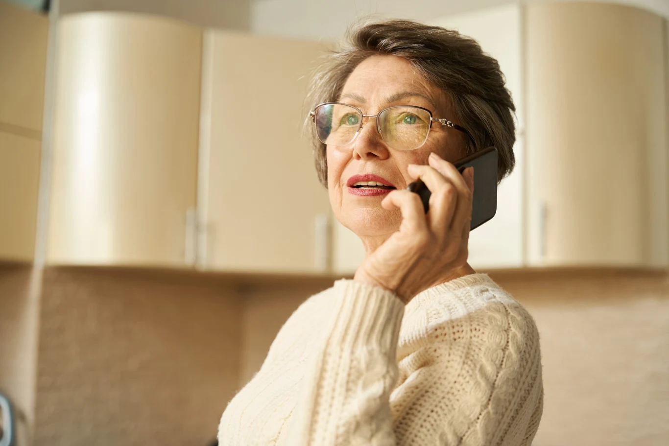 Koziorożec i Strzelec dowiedzą się czegoś, co postawi bliskich w nowym świetle Starsza kobieta w okularach, ubrana w jasny sweter, rozmawia przez telefon komórkowy w kuchni. Tło stanowią jasne szafki kuchenne.