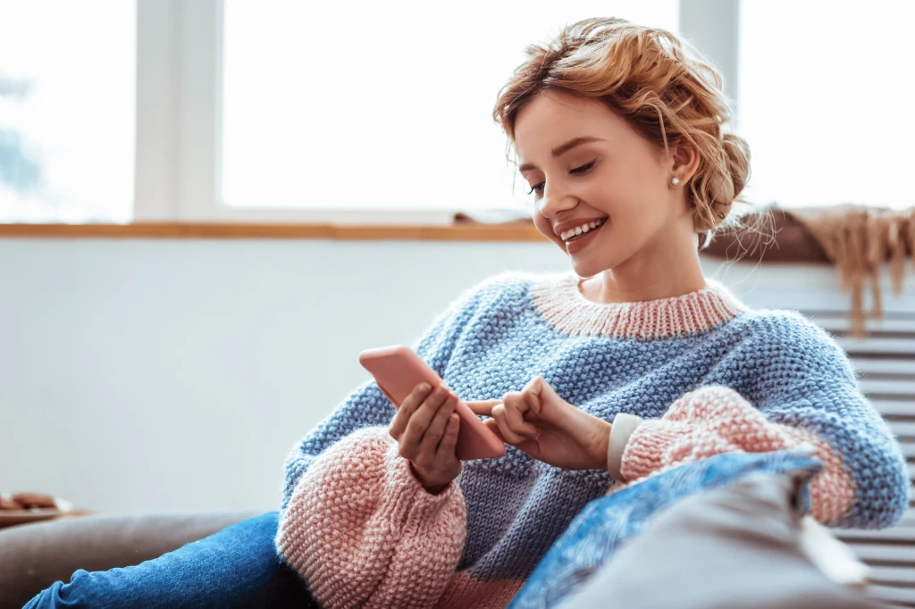 Uśmiechnięta młoda kobieta w pastelowym swetrze siedzi wygodnie na kanapie i korzysta ze smartfona, wokół panuje ciepła, domowa atmosfera.