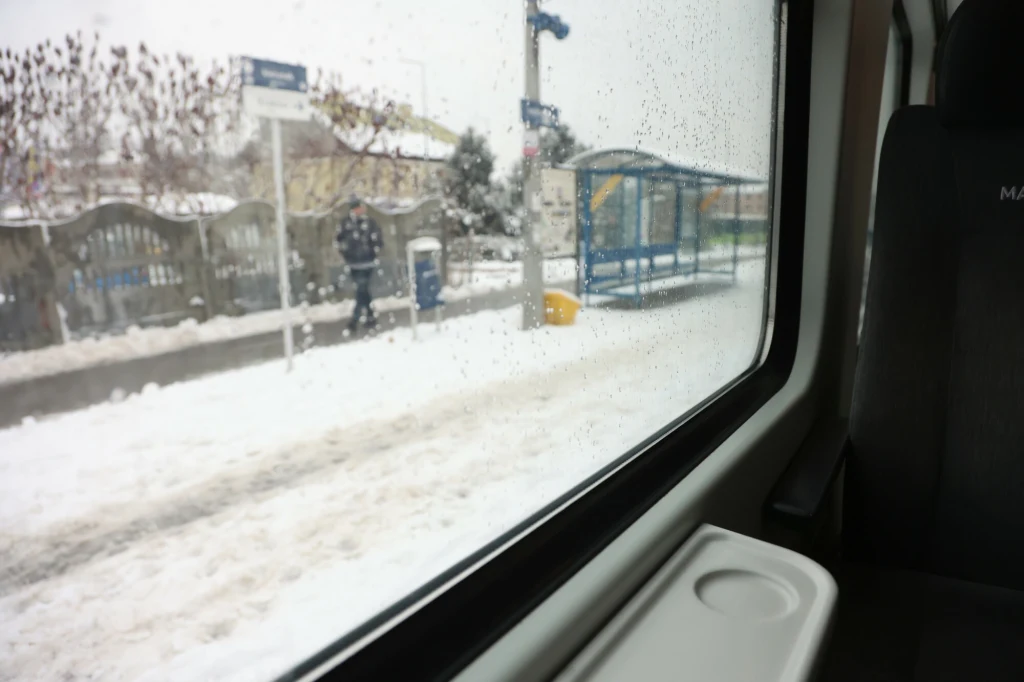 Chcesz mieć miejsce siedzące w pociągu? Lepiej kupi bilet z wyprzedzeniem Widok na przystanek autobusowy pokryty śniegiem, widziany przez zamgloną, wilgotną szybę wagonu. W tle stoi osoba ubrana zimowo, a cała scena utrzymana jest w zimowej, nieco zamglonej atmosferze z padającym śniegiem.