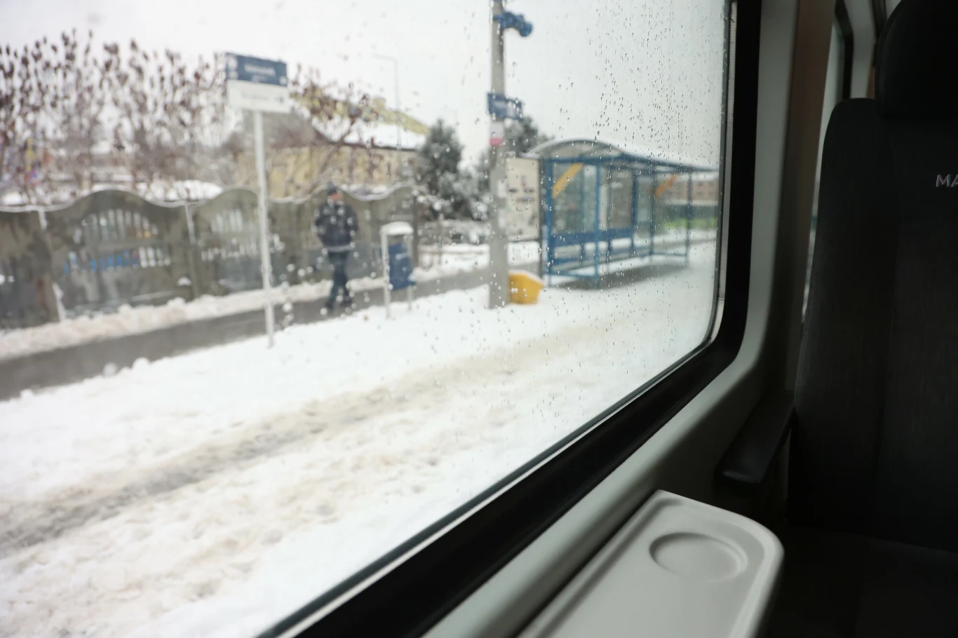 Widok na przystanek autobusowy pokryty śniegiem, widziany przez zamgloną, wilgotną szybę wagonu. W tle stoi osoba ubrana zimowo, a cała scena utrzymana jest w zimowej, nieco zamglonej atmosferze z padającym śniegiem.