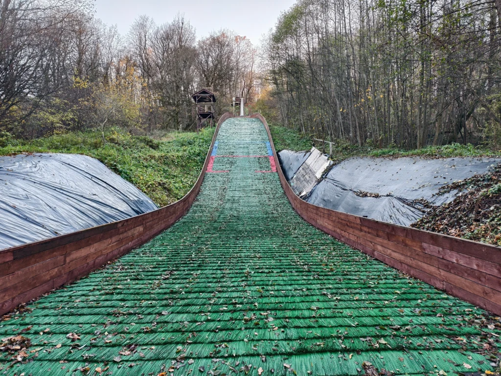Stroma, zielona skocznia narciarska otoczona drzewami w jesiennych barwach oraz metalowymi lub plastikowymi matami ochronnymi po bokach, w oddali drewniana wieża dla sędziów lub obserwatorów.