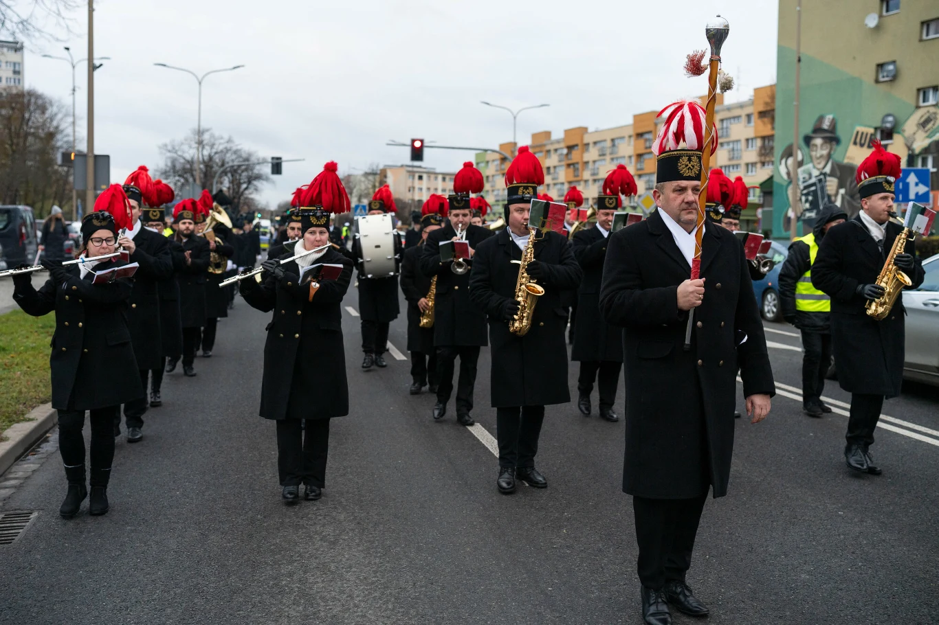 Parada to jeden ze stałych elementów obchodów Barbórki Orkiestra dęta maszeruje ulicą w odświętnych mundurach z ozdobnymi czapkami z czerwonymi pióropuszami, grając na instrumentach dętych i perkusyjnych, na tle bloków mieszkalnych i ulicznego ruchu.