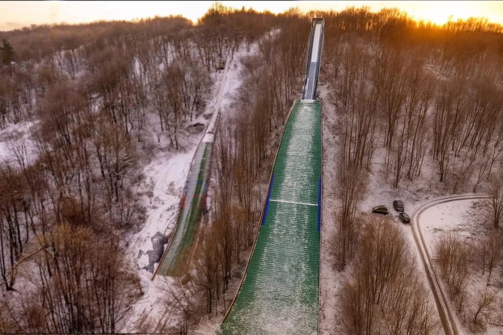 Zimowa skocznia narciarska otoczona przez zalesiony teren pokryty cienką warstwą śniegu, widoczne samochody zaparkowane przy drodze prowadzącej wśród drzew, słoneczny zachód tworzy ciepłe światło na tle zimowego krajobrazu.