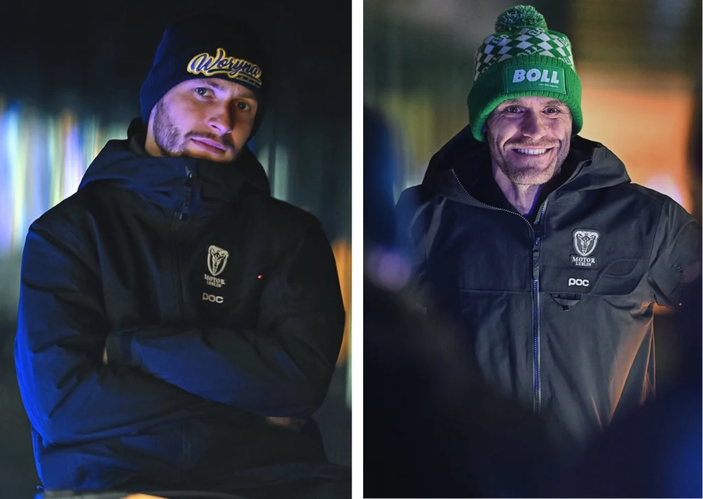 Dwóch mężczyzn pozujących w zimowych czapkach i czarnych kurtkach z wyeksponowanymi logotypami. Obaj stoją przed nieostrym, kolorowym tłem, jeden z nich patrzy z lekkim uśmiechem, drugi ma poważniejszy wyraz twarzy.