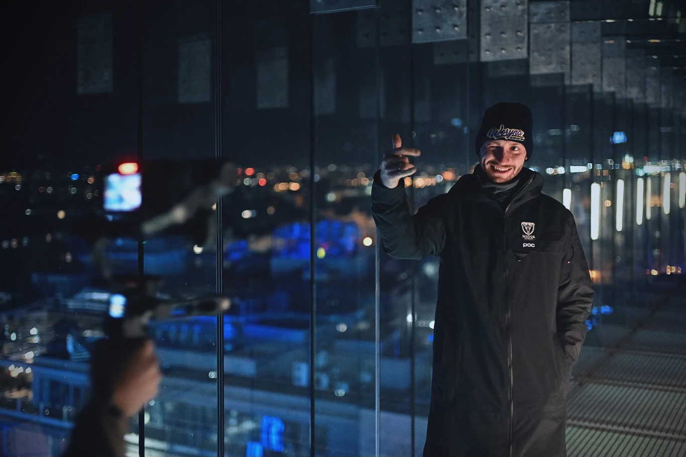 Mężczyzna w czarnej kurtce i czapce z uśmiechem pozuje przed kamerą na tle przeszklonego tarasu widokowego nocą z panoramą miasta rozświetloną światłami.