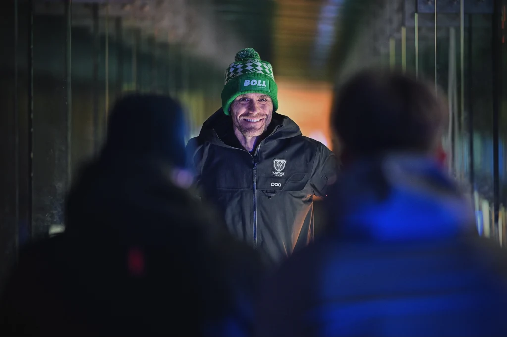 Mężczyzna w zielonej czapce i czarnej kurtce z logo drużyny żużlowej stoi uśmiechnięty w tunelu, naprzeciwko dwóch osób, których sylwetki są lekko rozmyte na pierwszym planie.