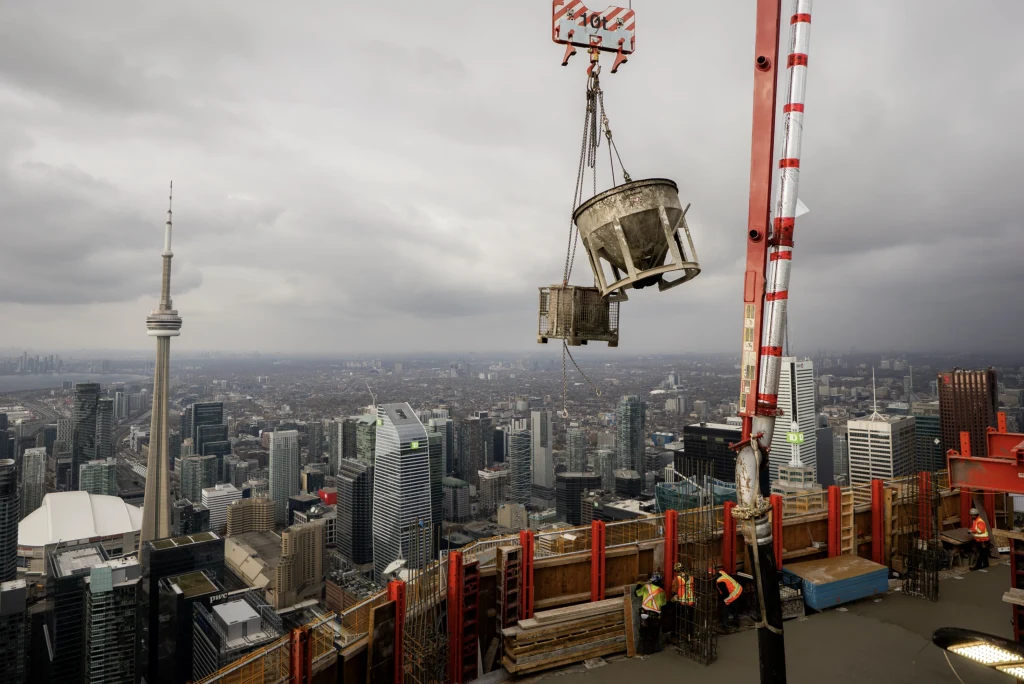 Z wieży widokowej Pinnacle SkyTower roztacza się widok na jezioro Ontario i panoramę Toronto, w tym słynną wieżę CN Tower Dźwig transportuje betonowy pojemnik nad szczytem wieżowca w centrum miasta, w tle liczne wieżowce i charakterystyczna wieża CN Tower, na pierwszym planie widoczni pracownicy budowy i elementy konstrukcyjne.