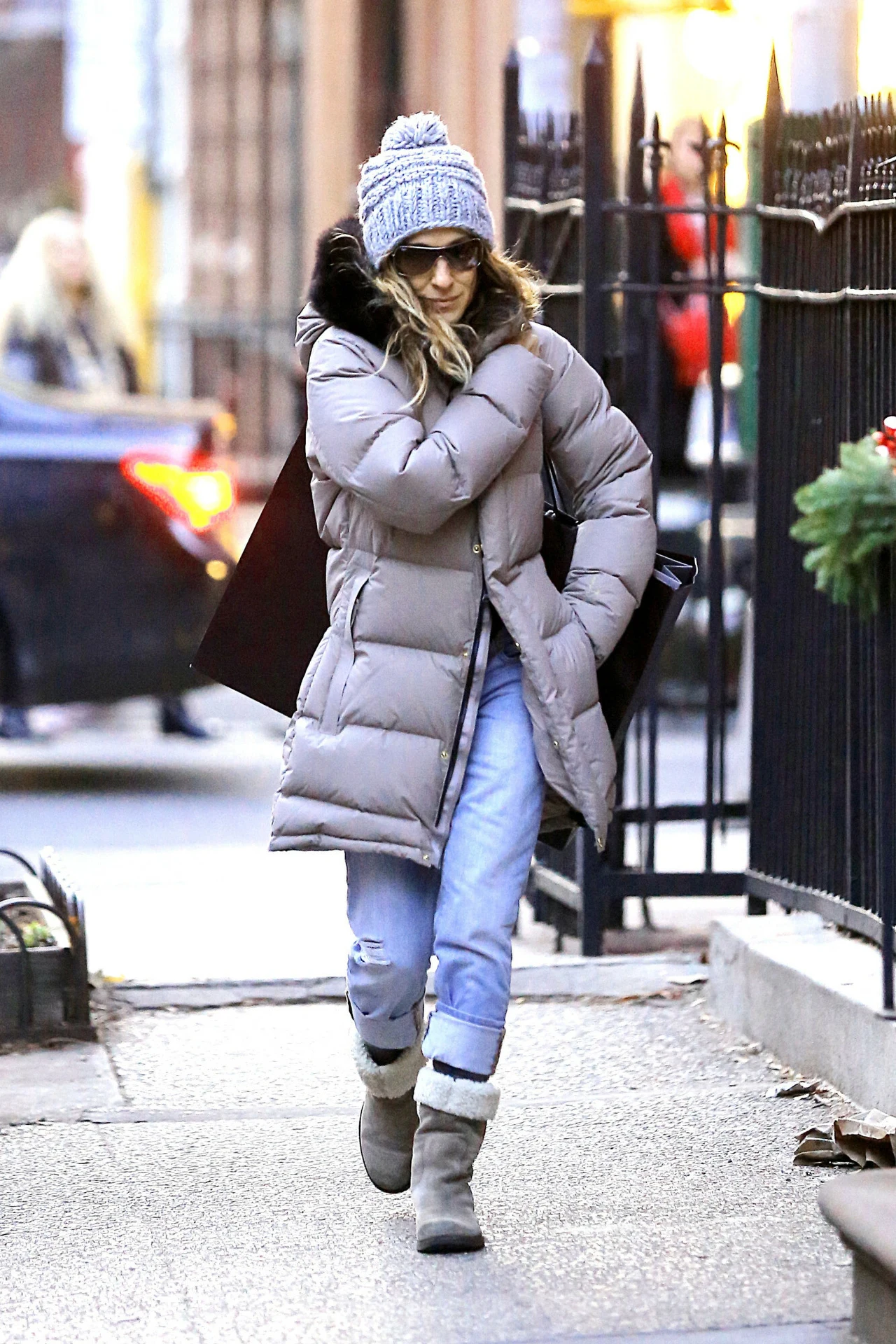 Kobieta ubrana w zimową kurtkę puchową, jasne jeansy, szare buty i czapkę z pomponem idzie chodnikiem w miejskiej przestrzeni. Trzyma ręce skrzyżowane na piersi, najprawdopodobniej z powodu zimna. W tle widoczne są ogrodzenie, fragment samochodu oraz d...