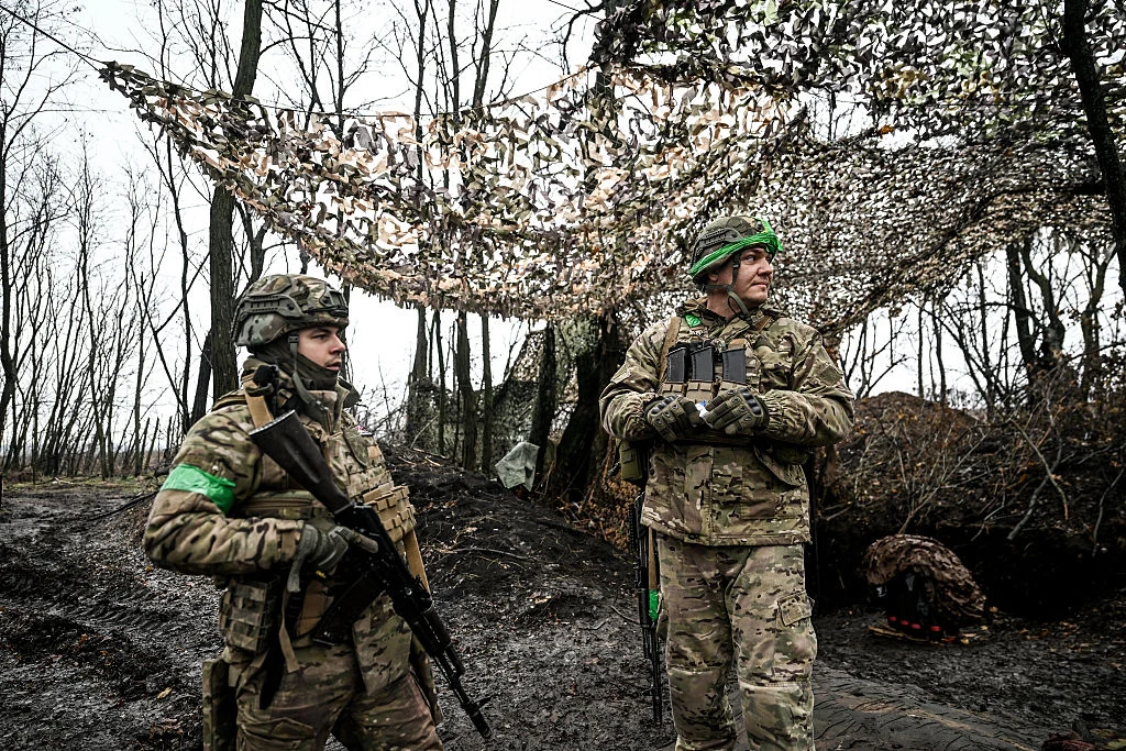 Ukraińscy żołnierze Dwóch żołnierzy w mundurach kamuflażowych z zielonymi opaskami na rękawach stoi na tle pozycji umocnionej siatką maskującą na terenie leśnym. Jeden z nich trzyma karabin, drugi obserwuje teren przez lornetkę.