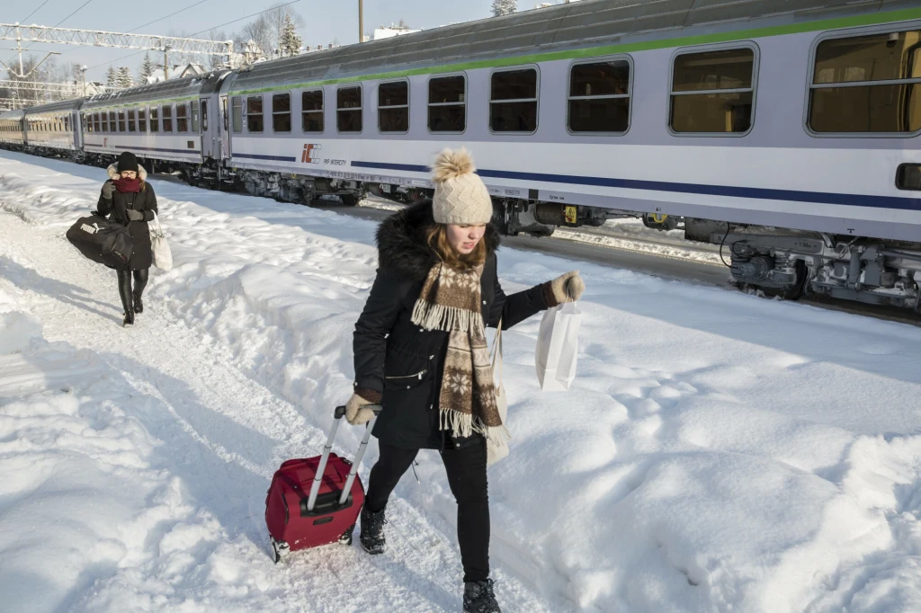 Bilety na pociągi w okresie świąt mogą szybko się wyprzedać Kobieta ciągnąca czerwoną walizkę po śniegu obok torów kolejowych, w tle wagon pociągu pasażerskiego i druga osoba idąca z białą torbą. Mroźna, zimowa aura i wysoka warstwa śniegu na peronie.