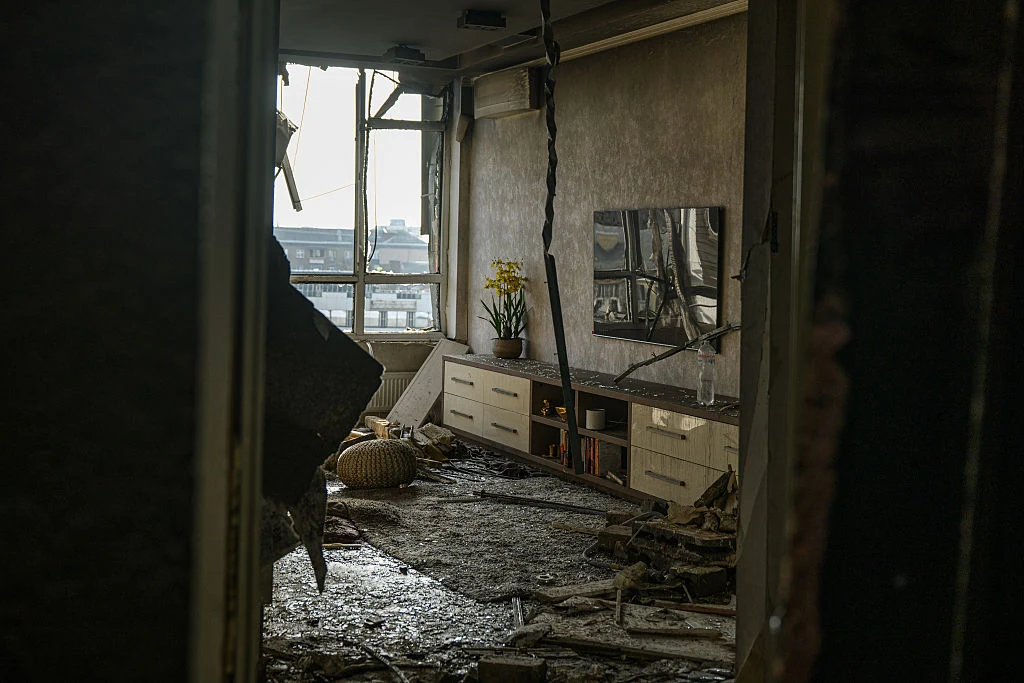 Pokój dzienny z wybitymi oknami, uszkodzonym sufitem, roztrzaskanymi meblami i fragmentami zniszczonych ścian, na podłodze leżą gruz i błoto, a w tle widać zabrudzone miasto przez powybijane szyby.