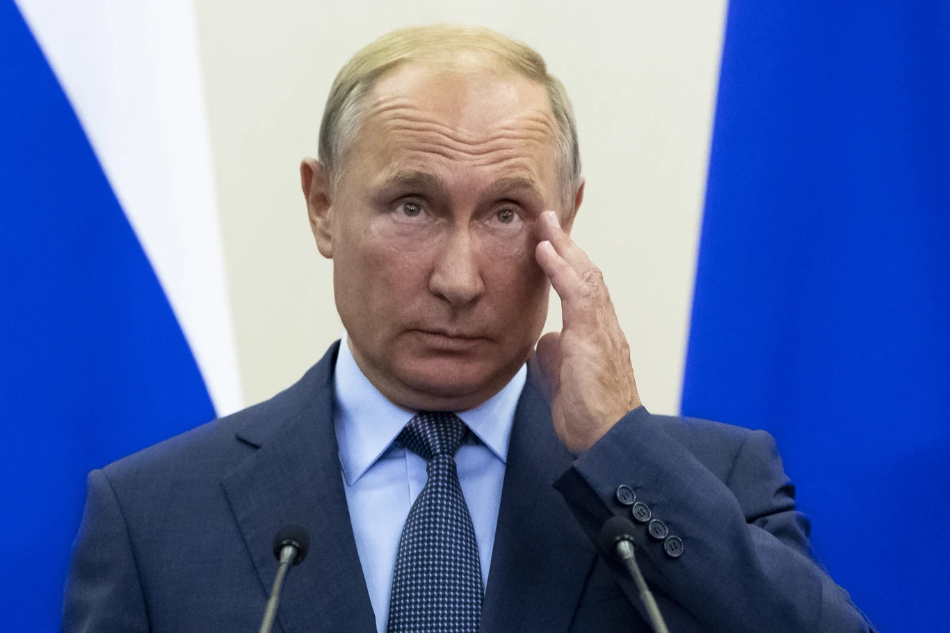 Prezydent Rosji Władimir Putin Mężczyzna w garniturze stoi na tle niebieskich i białych flag, trzyma rękę przy twarzy, wyglądając poważnie.