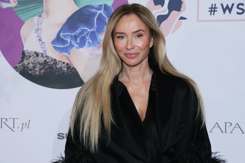 Kobieta z długimi blond włosami ubrana w czarną stylową marynarkę pozuje na tle kolorowej ścianki z graficznymi motywami i logotypami.