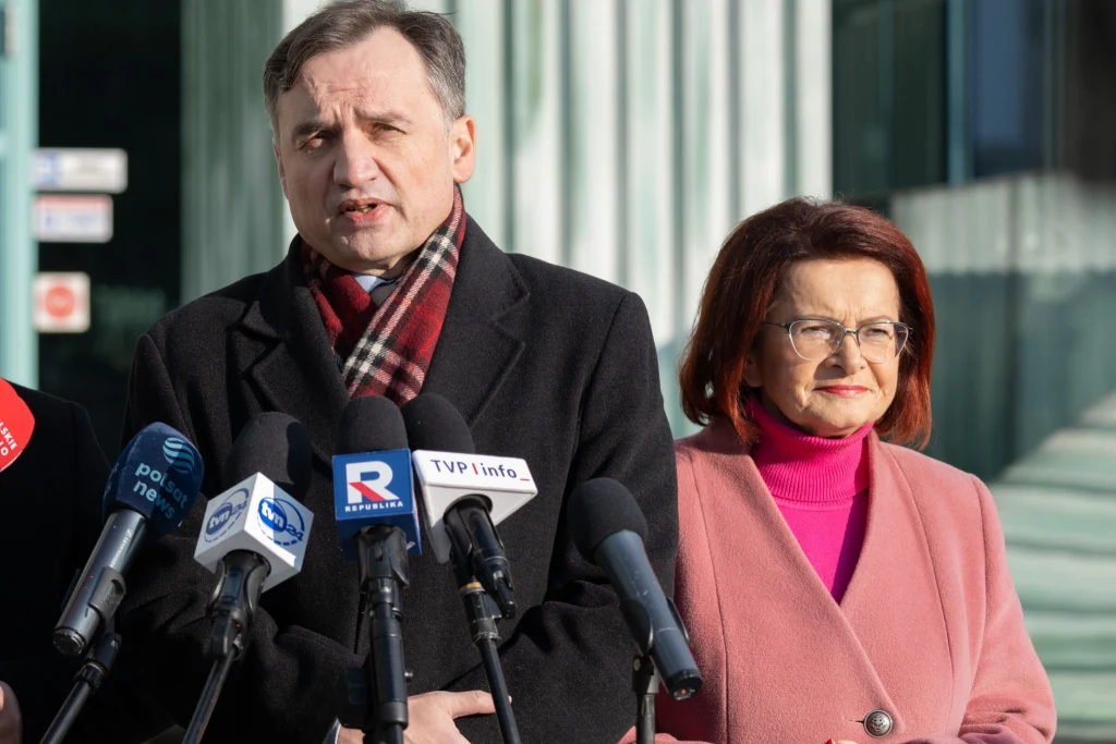 W sprawie Funduszu Sprawiedliwości pojawiło się nazwisko posłanki Marii Kurowskiej Mężczyzna w czarnym płaszczu i czerwonym szaliku przemawia do mikrofonów różnych stacji telewizyjnych podczas konferencji prasowej, obok niego kobieta w różowym płaszczu i okularach na tle nowoczesnego budynku.