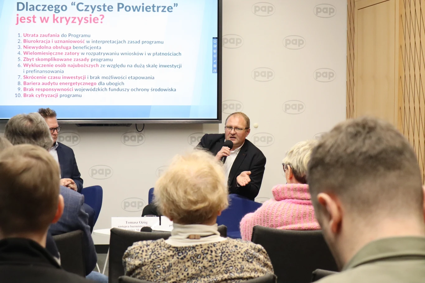 - Dostęp do nowej wersji programu "Czyste Powietrze" dla osób o niskich dochodach jest bardzo ograniczony - zauważa Tomasz Ożóg, z-ca burmistrza Skawiny Publiczna debata lub konferencja z udziałem eksperta omawiającego kryzys programu 'Czyste Powietrze' przed grupą słuchaczy, na ekranie za prelegentem znajduje się lista punktów dotyczących przyczyn kryzysu.