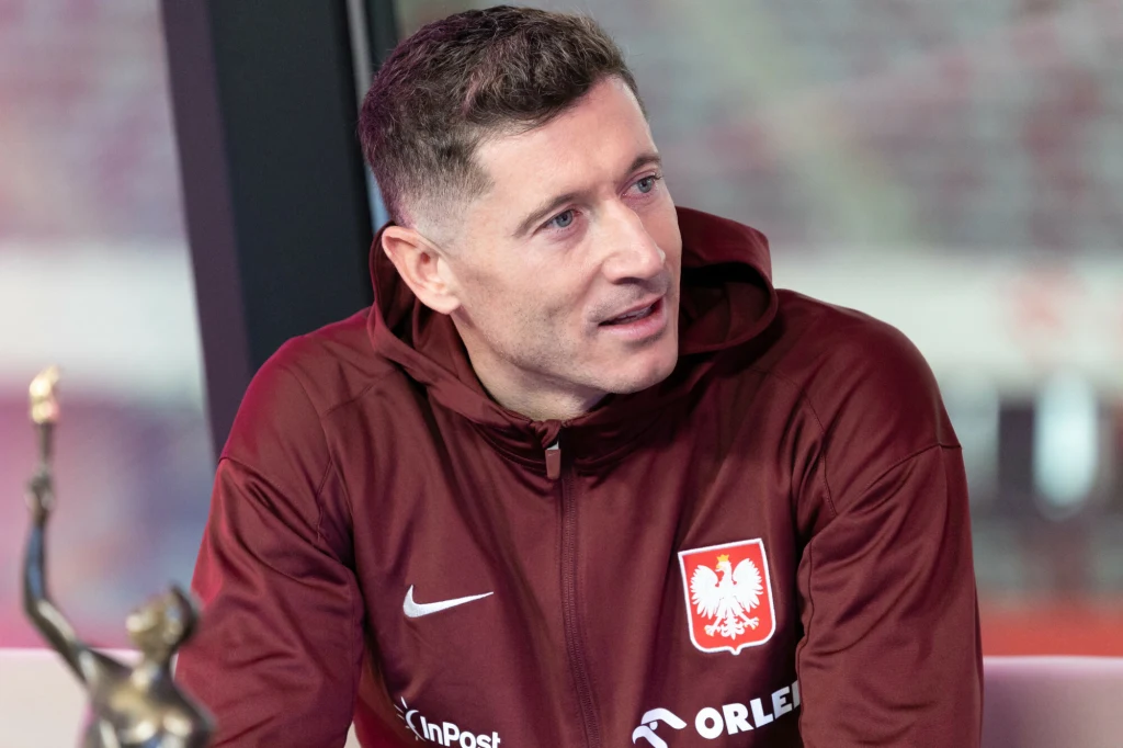 Mężczyzna w bordowej bluzie reprezentacji Polski z orłem na piersi, siedzi i rozmawia, w tle rozmyte światło. Po lewej stronie widoczna statuetka.