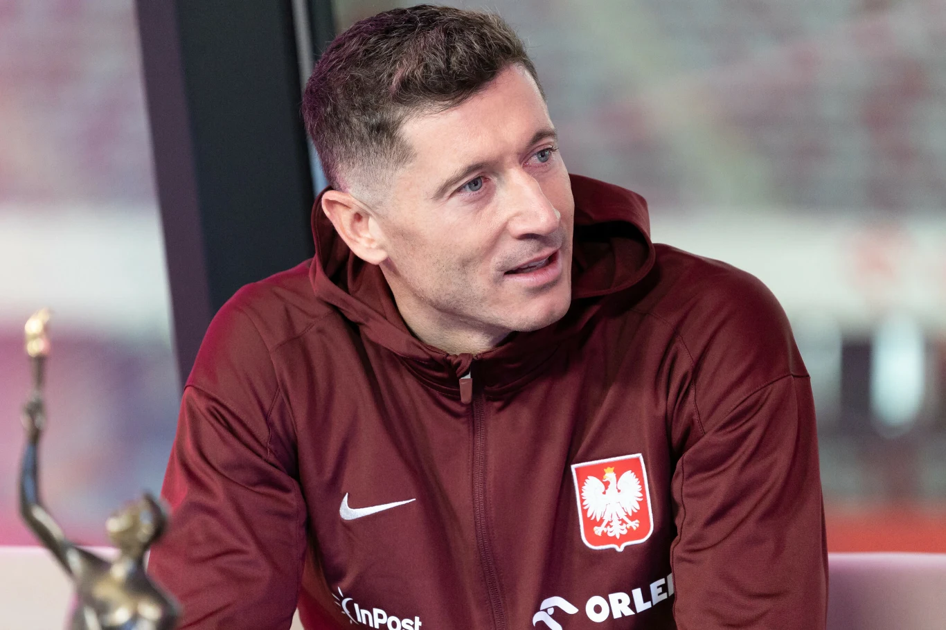 Mężczyzna w bordowej bluzie reprezentacji Polski z orłem na piersi, siedzi i rozmawia, w tle rozmyte światło. Po lewej stronie widoczna statuetka.