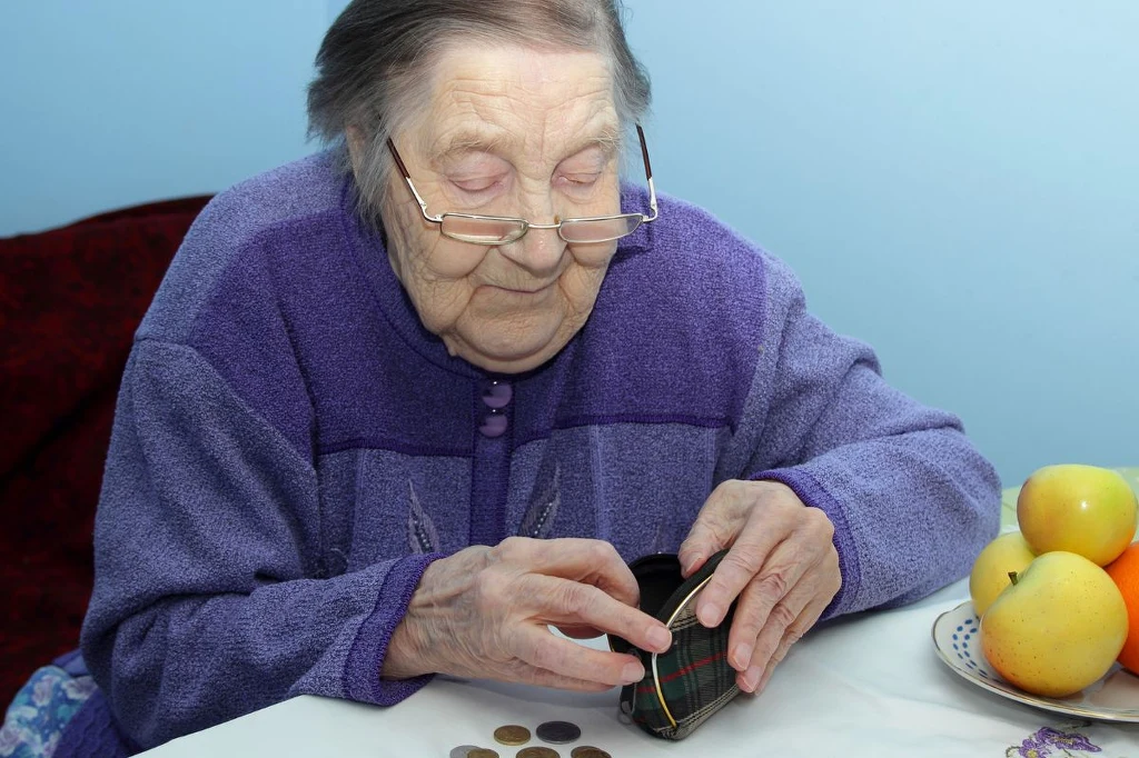 W grudniu zmienią się terminy wypłat emerytur. Niektórzy otrzymają dwa przelewy. Starsza kobieta w okularach liczy monety, trzymając w ręku portmonetkę. Na stole znajduje się kilka monet oraz talerz z żółtymi jabłkami.