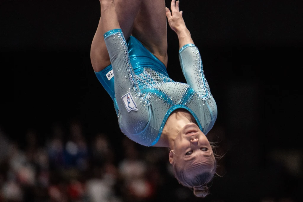 Angielina Mielnikowa Zawodniczka gimnastyki sportowej w błękitnym stroju wykonująca skomplikowaną figurę akrobatyczną w powietrzu na tle rozmazanej publiczności.