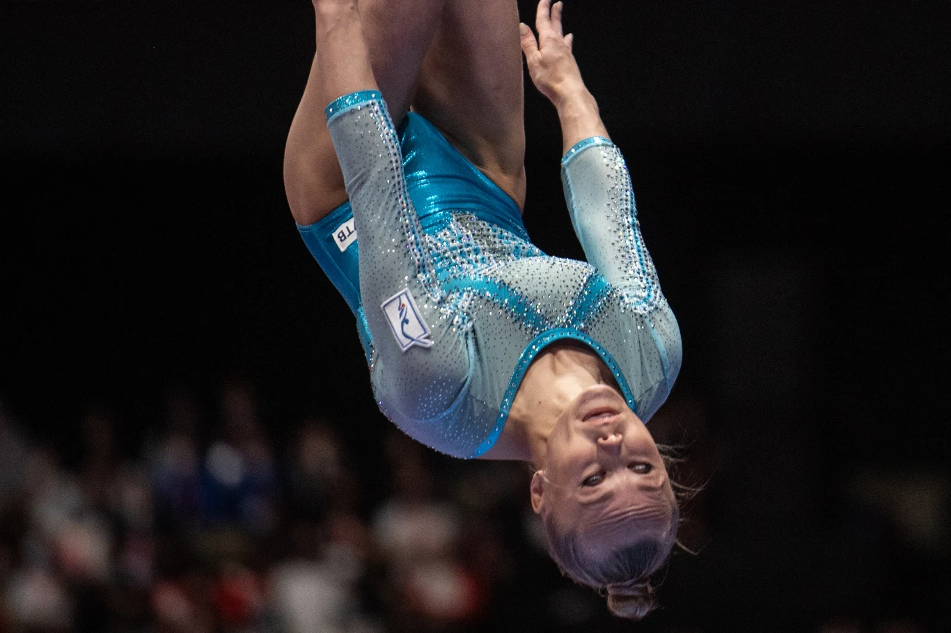Zawodniczka gimnastyki sportowej w błękitnym stroju wykonująca skomplikowaną figurę akrobatyczną w powietrzu na tle rozmazanej publiczności.