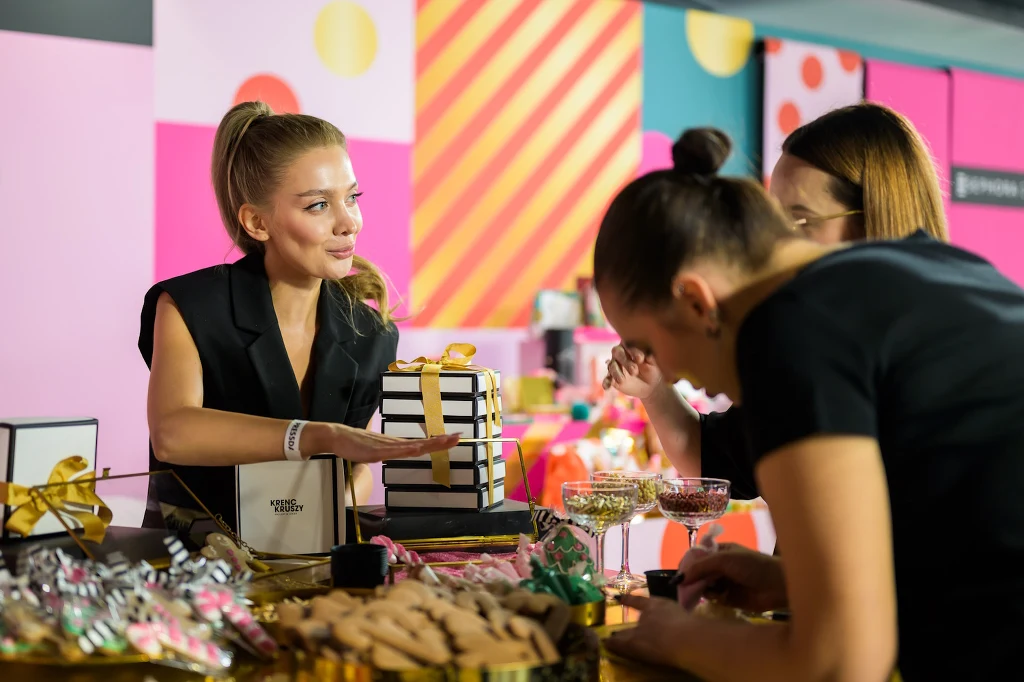Młoda kobieta w czarnym ubraniu rozmawia z dwiema innymi kobietami przy udekorowanym stole z prezentami, słodyczami i kieliszkami z napojami w jasnym, kolorowym wnętrzu.