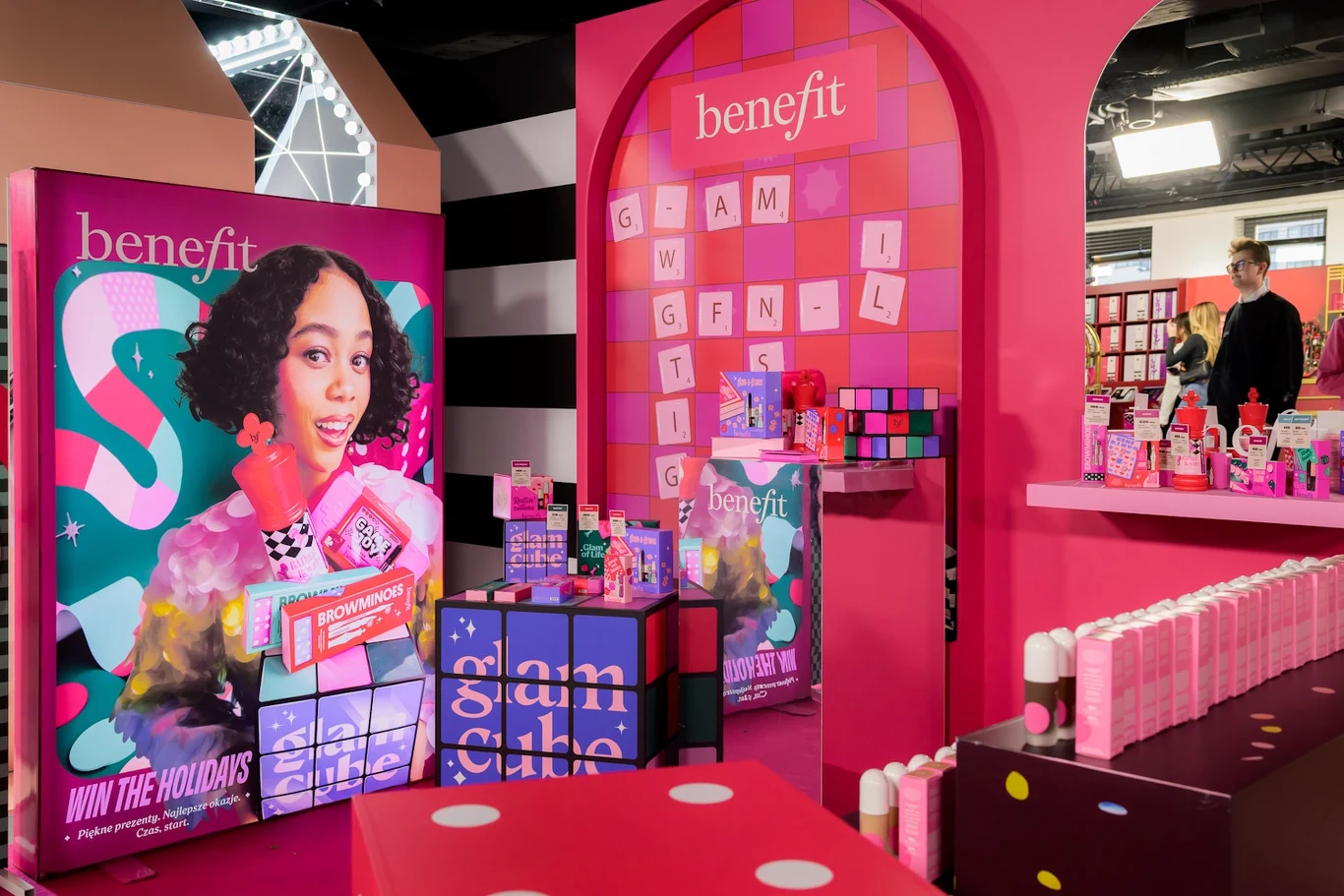 Kolorowe stoisko promocyjne marki Benefit, pełne opakowań kosmetyków ustawionych na stolikach oraz dużych plakatów reklamowych z wizerunkiem młodej kobiety. Tło utrzymane w intensywnie różowej kolorystyce, wystrój ozdobiony motywami reklamowymi i nazwą...