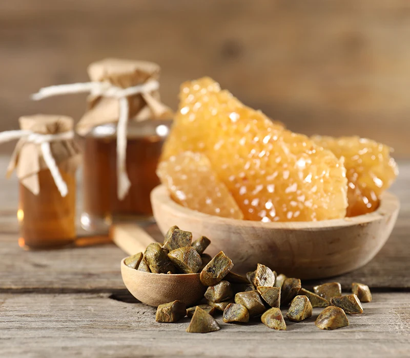 Kawałki propolisu w drewnianej łyżce i miseczce, plastry miodu oraz słoiczki z miodem na drewnianym tle.