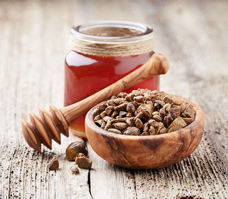 Drewniana miska wypełniona propolisem, obok znajduje się baryłka miodu oraz pałeczka do miodu, całość ustawiona na rustykalnym, drewnianym stole.