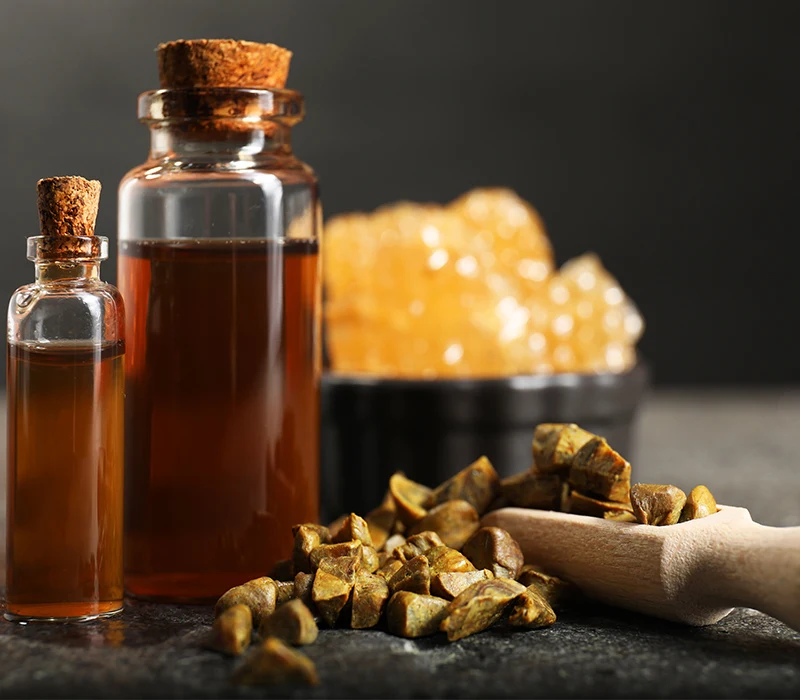 Butelki z brązowym płynem z korkiem, kawałki propolisu na stole oraz fragment plastra miodu w tle.