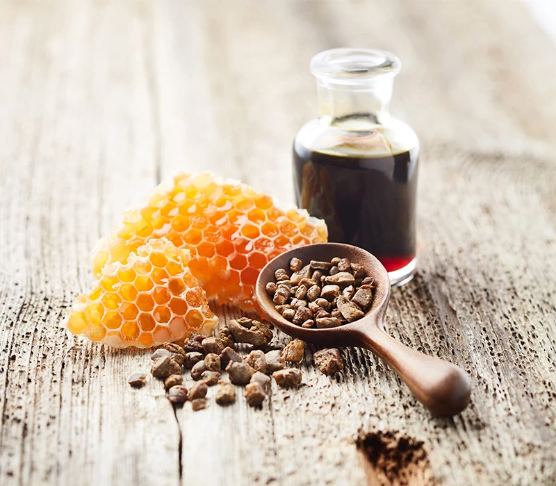 Plastry miodu, nasiona ostropestu w drewnianej łyżce oraz butelka z ciemnym płynem ustawione na drewnianym stole.