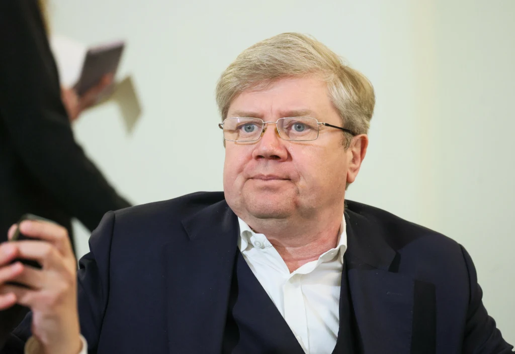 Mężczyzna w okularach i eleganckiej marynarce wpatruje się przed siebie, wykonując gest ręką, w tle niewyraźna postać trzymająca notes lub dokumenty.