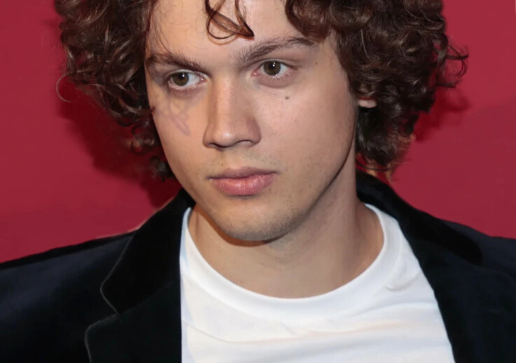 Nataniel Pągowski Młody mężczyzna o kręconych brązowych włosach, ubrany w biały t-shirt i ciemną marynarkę, stoi na tle jednolitej czerwonej ściany, patrząc lekko w bok z neutralnym wyrazem twarzy.