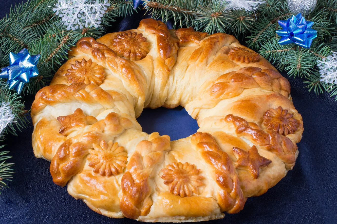 Serowy wieniec będzie doskonałą przekąską na święta. Zrobisz go błyskawicznie Okrągłe ciasto drożdżowe z ozdobami w kształcie liści i kwiatów, otoczone zielonymi gałązkami świerku udekorowanymi niebieskimi kokardami i śnieżynkami.