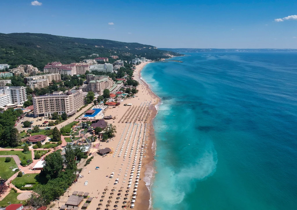 Rozległa, piaszczysta plaża z równo rozstawionymi parasolami i leżakami, granicząca z błękitnym morzem oraz otoczona szeregiem hoteli i budynków wypoczynkowych zadrzewionych wzgórz w tle.