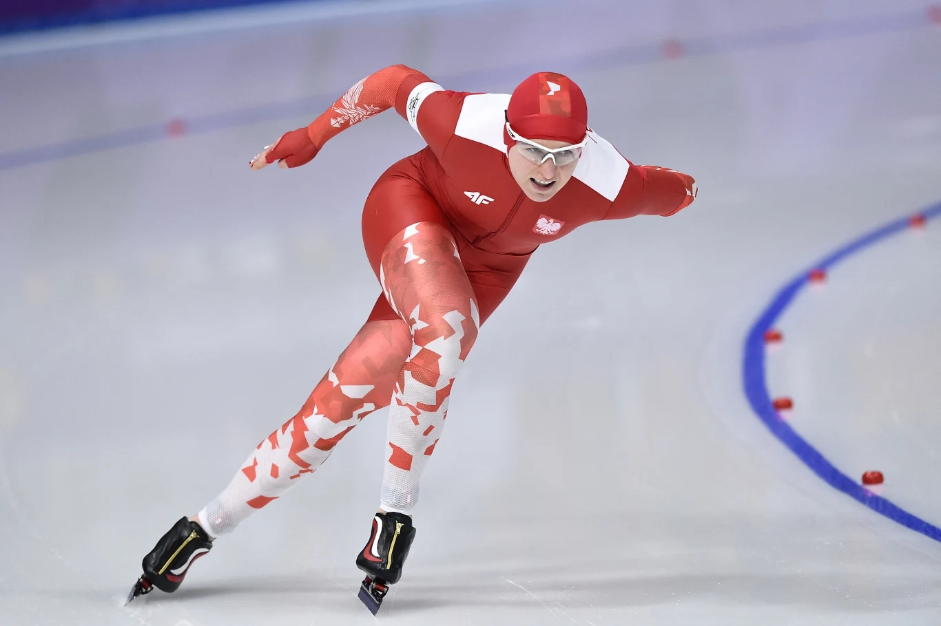Łyżwiarz szybki w czerwonym stroju z biało-czerwonym motywem w trakcie wyścigu na lodowisku, dynamiczny ruch, wyraz skupienia na twarzy, wokół tor oznaczony niebieskimi liniami i czerwonymi punktami.