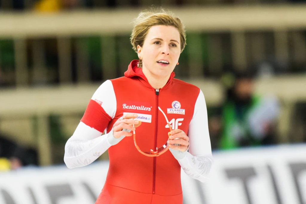 Zawodniczka w czerwono-białym stroju sportowym na lodowisku, trzymająca w rękach okulary ochronne, skupiona i skoncentrowana podczas startu w zawodach łyżwiarskich.