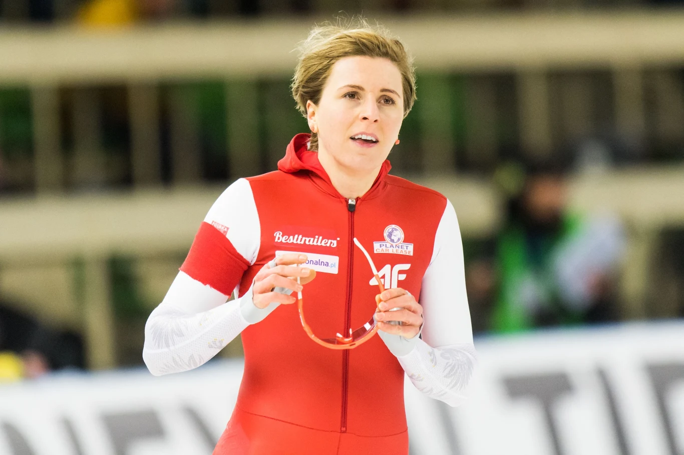 Zawodniczka w czerwono-białym stroju sportowym na lodowisku, trzymająca w rękach okulary ochronne, skupiona i skoncentrowana podczas startu w zawodach łyżwiarskich.