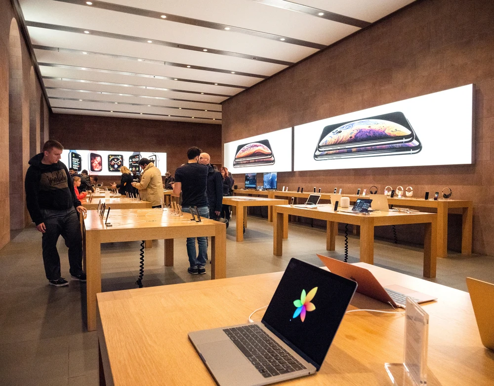 Nowoczesne wnętrze sklepu z elektroniką, na pierwszym planie drewniane stoły z wystawionymi laptopami, w tle klienci oglądający urządzenia i duże podświetlane reklamy smartfonów na ścianach.