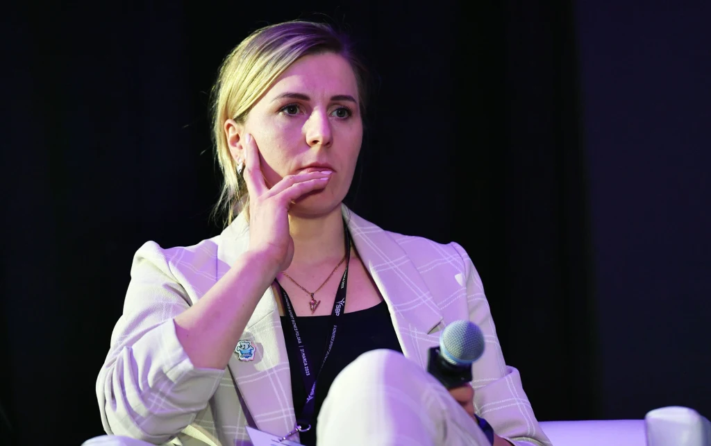 Kobieta w jasnej marynarce siedzi na scenie z mikrofonem w dłoni, spogląda poważnie w bok, dotykając twarzy palcami, w tle ciemne tło.