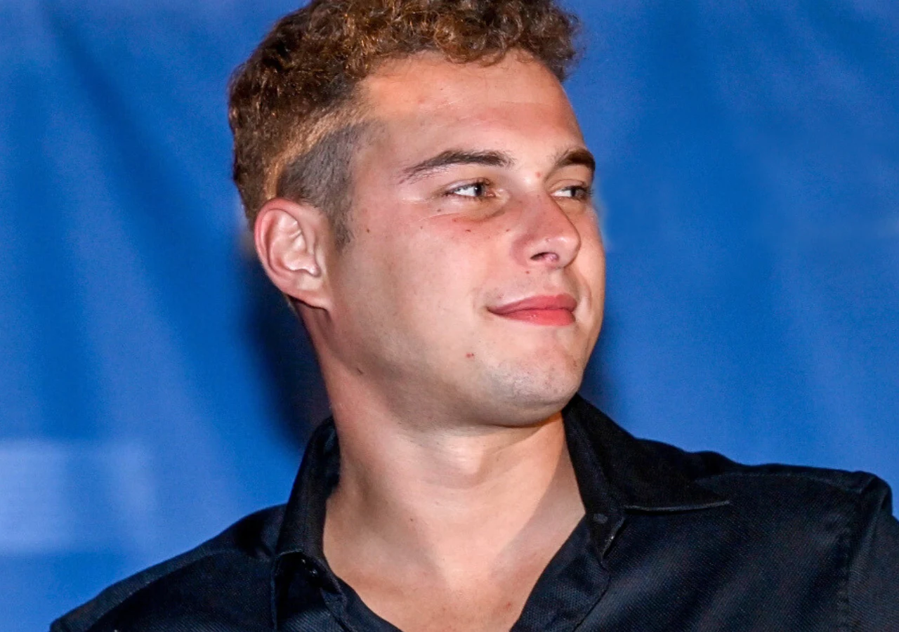 Adam Zdrójkowski Młody mężczyzna z krótkimi, kręconymi włosami i lekkim zarostem ubrany w elegancką, czarną koszulę spogląda w bok na tle niebieskiej, rozmytej scenerii.