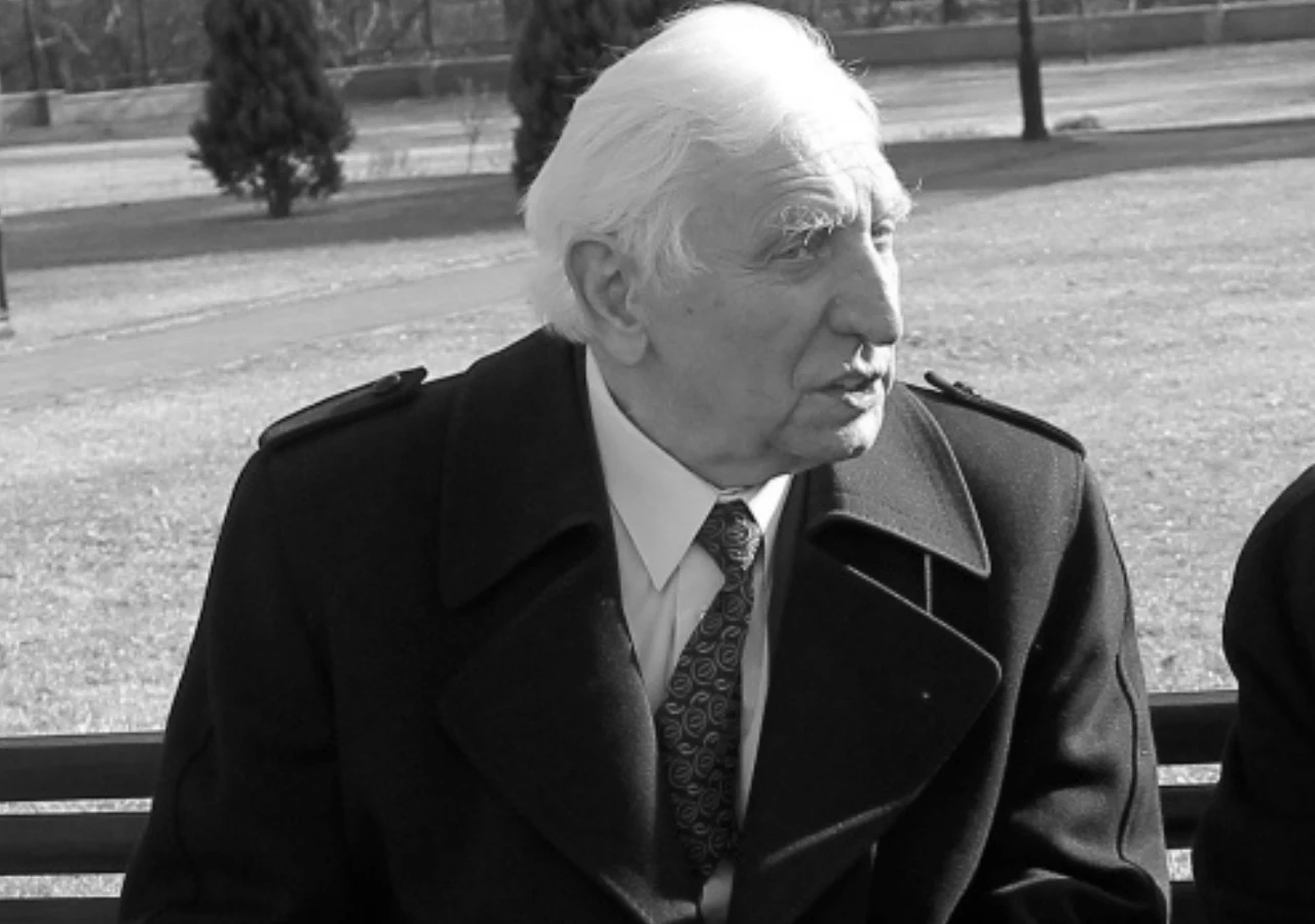 Stanisław Sparażyński nie żyje Starszy mężczyzna z siwymi włosami ubrany w elegancki płaszcz i krawat siedzi na ławce w parku, spoglądając w bok. Tło stanowi trawnik i drzewa, kadrowanie skupia się głównie na twarzy i sylwetce mężczyzny.