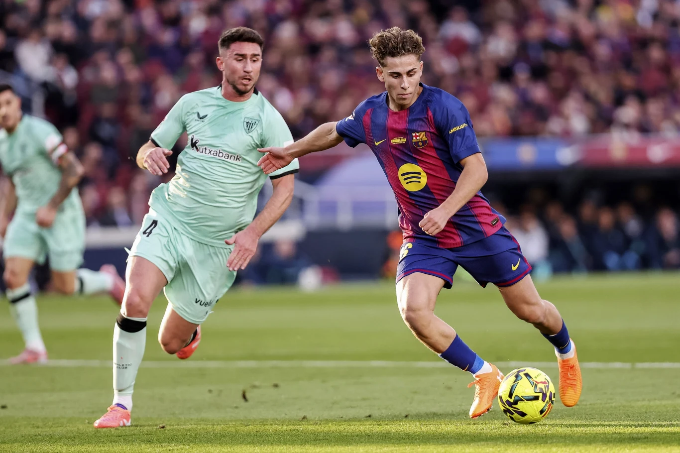 Dwóch piłkarzy walczy o piłkę podczas meczu piłki nożnej na stadionie, widoczna intensywna rywalizacja sportowa, piłkarz w stroju Barcelony kontroluje piłkę, a zawodnik drużyny przeciwnej próbuje go zatrzymać.