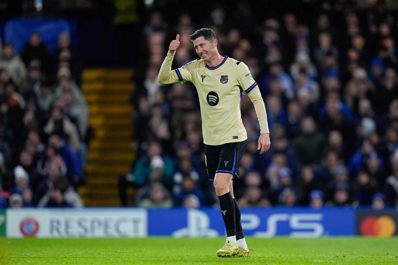 Piłkarz ubrany w jasny strój drużyny z emblematem Spotify na piersi pokazuje gest uniesionego kciuka na tle tłumu kibiców na stadionie. W tle widoczna trybuna oraz rozmyta publiczność.