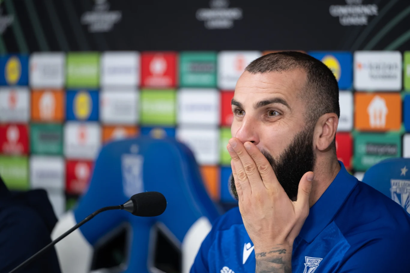 Mikael Ishak przed meczem w Lidze Konferencji Mężczyzna z brodą w niebieskiej koszulce klubu sportowego siedzi przy mikrofonie na tle ściany z kolorowymi logotypami sponsorów i zakrywa usta dłonią, wyrażając zaskoczenie lub emocje.